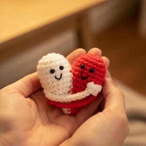 Può includere: Un cuore all'uncinetto fatto a mano, rosso e bianco, con facce sorridenti e braccia che si abbracciano. Le due metà del cuore sono tenute in mani aperte, mostrando l'artigianato e il design. Il cuore è un simbolo d'amore.