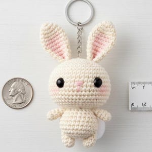 May include: A handmade crochet bunny keychain in cream with pink accents on the ears and cheeks. The bunny has black eyes and a small white tail. A quarter and a ruler are next to the bunny for size reference.