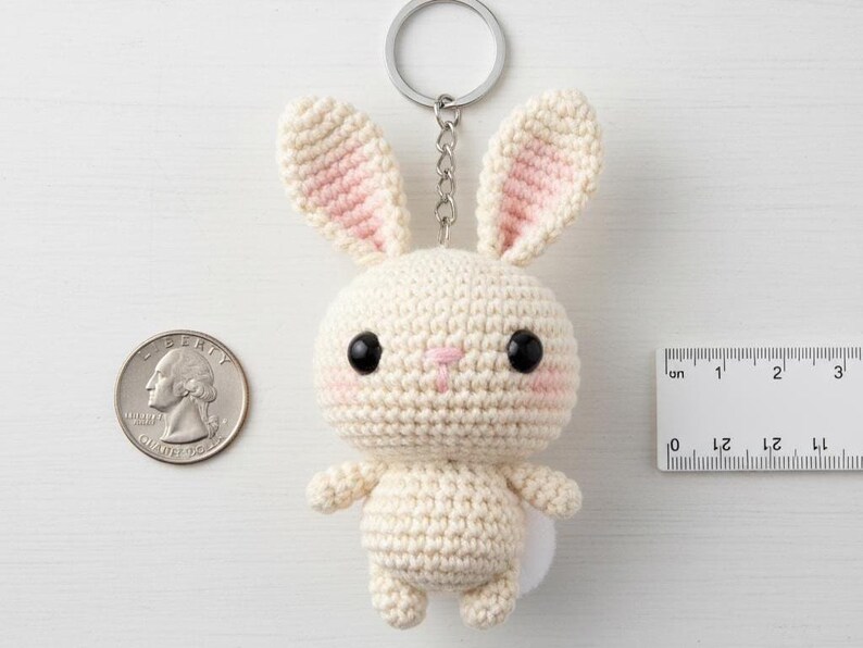 May include: A handmade crochet bunny keychain in cream with pink accents on the ears and cheeks. The bunny has black eyes and a small white tail. A quarter and a ruler are next to the bunny for size reference.