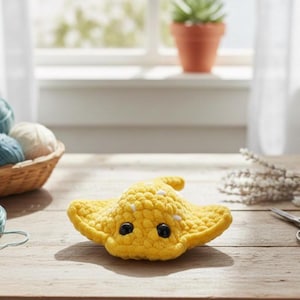 May include: A handmade yellow crochet stingray with black eyes and white accents sits on a wooden surface. A basket of yarn balls in various shades of blue and white, a ball of teal yarn, and scissors are also on the table. The background includes a window and a small potted plant.