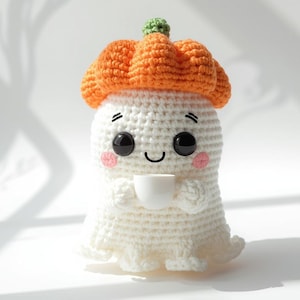Könnte beinhalten: Handgemachte gehäkelte Geisterpuppe mit orangefarbenem Kürbishut und einer kleinen weißen Tasse. Der Geist hat schwarze Augen, rosige Wangen und ein lächelndes Gesicht. Die Puppe ist aus weißem Garn.