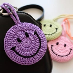 Puede incluir: Un llavero de carita sonriente morado tejido a ganchillo, sujeto a un bolso negro. Varios llaveros de carita sonriente tejidos a ganchillo en colores pastel son visibles en el fondo.