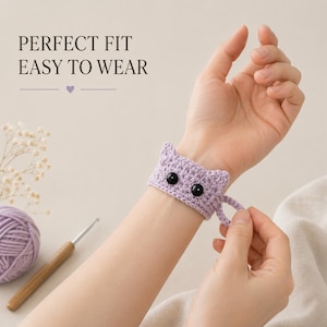 May include: A light purple crocheted wristband featuring a cat face design with two pointed ears and two round black bead eyes. The text PERFECT FIT EASY TO WEAR is visible in the upper left corner. The wristband has a small loop closure.