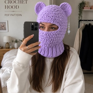 May include: A light purple crochet balaclava hood featuring two rounded ears on top and a ribbed neck section. A dangling charm with beads and a star hangs from the right ear. Text on the image reads TRENDING CROCHET HOOD PDF PATTERN.