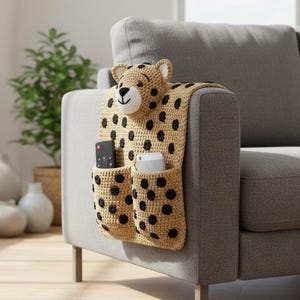 Puede incluir: Un organizador de brazo de sofá tejido en beige y negro con forma de guepardo. El organizador tiene dos bolsillos y una cabeza de guepardo en la parte superior. Un mando a distancia y un teléfono están en los bolsillos. El organizador está en un sofá gris.