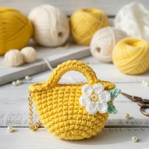May include: A handmade yellow crochet bag with a white flower and green leaves attached. The bag has a small handle and a gold chain. The background includes balls of yellow and cream-colored yarn, pearls, and scissors.