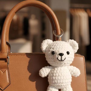 Könnte beinhalten: Ein weißer gehäkelter Teddybär-Schlüsselanhänger mit einem schwarz gestickten Gesicht, befestigt an einer braunen Lederhandtasche. Der Bär hat kleine Gliedmaßen und ist aus weichem, strukturiertem Garn gefertigt. Die Handtasche hat einen braunen Griff.