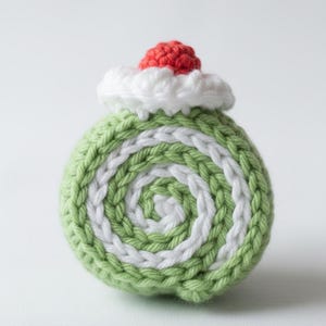 May include: A crocheted ornament in the shape of a dessert. It has a spiral pattern of green and white yarn. A red crocheted cherry sits on top of a white, fluffy, crocheted cloud, creating a whimsical design.