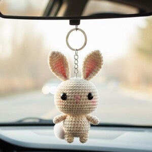 May include: A crocheted bunny car charm hanging from a rearview mirror. The bunny is beige with pink accents on the ears and cheeks, and a small white pom-pom tail. It has black eyes and is suspended by a silver chain.