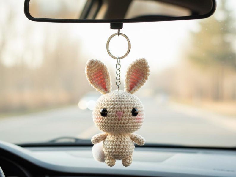 May include: A crocheted bunny car charm hanging from a rearview mirror. The bunny is beige with pink accents on the ears and cheeks, and a small white pom-pom tail. It has black eyes and is suspended by a silver chain.