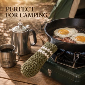 May include: A dark olive green crochet handle cover with two white horizontal stripes, fitted onto the handle of a black cast iron skillet containing fried eggs and bacon, alongside a stainless steel coffee pot and a metal mug.