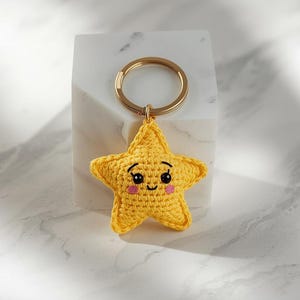 Può includere: Un portachiavi a forma di stella gialla all'uncinetto con un anello color oro. La stella ha una faccia sorridente con occhi neri e guance rosa. Il portachiavi è esposto su un blocco bianco su sfondo marmoreo.