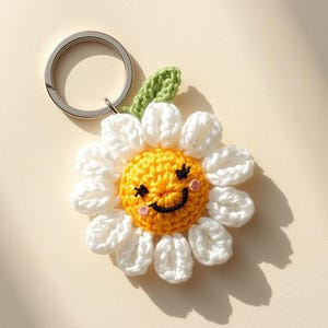 May include: A handmade crochet daisy keychain with a smiling face. The daisy has white petals, a yellow center, and a green leaf. The keychain has a silver metal ring.