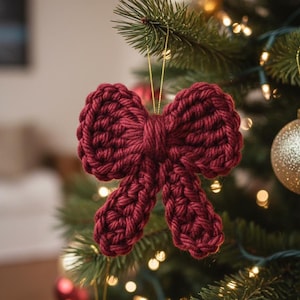 Pode incluir: Um enfeite de Natal de laço de crochê bordô pendurado em uma árvore de Natal decorada. O laço é feito de fio grosso e tem uma aparência texturizada. A árvore é adornada com luzes e outros enfeites.