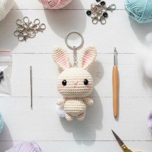 May include: A handmade crochet bunny keychain with pink accents, surrounded by crafting supplies like yarn, a crochet hook, and scissors. The bunny has a white body, pink ears, and a fluffy white tail. The scene is set on a white wooden surface.