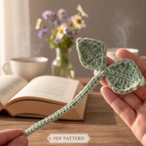 Op de afbeelding: Een handgemaakte, lichtgroene gehaakte bladwijzer in de vorm van bladeren. De bladwijzer wordt in een hand gehouden, met een open boek en een kop koffie op de achtergrond. De tekst "PDF PATTERN" staat onderaan.
