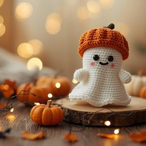 May include: A crocheted ghost figure wearing an orange pumpkin hat, set on a wooden slice. The ghost has a smiling face and is surrounded by miniature pumpkins, fall leaves, and string lights, creating a festive autumn scene.