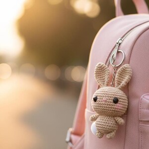 May include: A crocheted bunny keychain in tan and white hangs from a pink backpack. The bunny has black eyes, pink cheeks, and a small white tail. The backpack has a silver zipper and a small pocket.