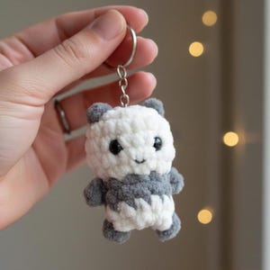 May include: A handmade crochet panda bear keychain. The panda is white and gray with black eyes and a small smile. The keychain is attached to a silver metal ring and chain. The panda is approximately 3 inches tall.