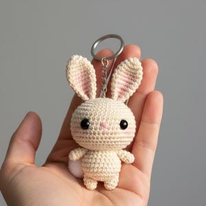 May include: A handmade crochet bunny keychain in cream with pink accents on the ears and nose. The bunny has large black eyes and a small white pom-pom tail. The keychain is held in a hand against a neutral background.