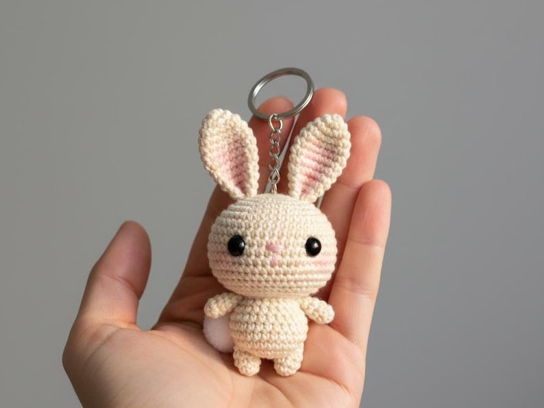 May include: A handmade crochet bunny keychain in cream with pink accents on the ears and nose. The bunny has large black eyes and a small white pom-pom tail. The keychain is held in a hand against a neutral background.