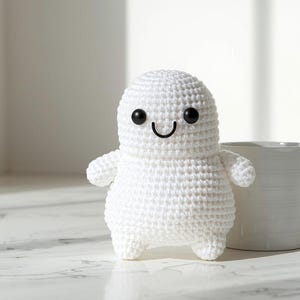 May include: A handmade white crochet ghost with black eyes and a smiling mouth. The ghost is standing next to a white mug on a white marble surface. The ghost is a cute, whimsical design.