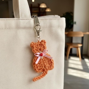 May include: Orange crocheted cat keychain with a pink ribbon bow and a small red bead. The cat is attached to a silver-colored metal clasp. The keychain is hanging from a white canvas tote bag.