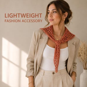 Puede incluir: Un pañuelo triangular de ganchillo color óxido con un patrón calado atado al cuello, sobre una camiseta blanca de canalé y una americana beige. El texto LIGHTWEIGHT FASHION ACCESSORY aparece impreso en letras color óxido en la parte superior.