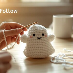 May include: A crocheted white ghost with a smiling face and small arms. The ghost is being crocheted with a gold crochet hook and white yarn. Text in the image reads "Easy To Follow Beginner Project".