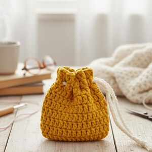 May include: A mustard yellow crochet drawstring pouch with a white drawstring. The pouch is on a light wooden surface, with a cup of coffee, books, and yarn in the background. The pouch is handmade.