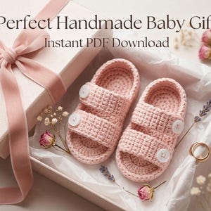 May include: A pair of handmade pink crochet baby sandals with white buttons, presented in a gift box with a pink ribbon. The box is lined with white paper and decorated with dried flowers. Text on the image reads: Perfect Handmade Baby Gift Instant PDF Download.