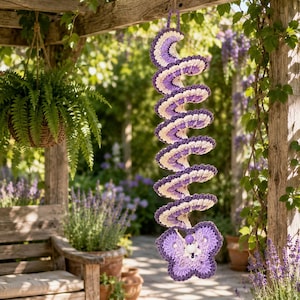May include: A spiral-shaped crochet hanging decoration featuring alternating bands of purple and cream yarn. The structure ends with a purple crocheted butterfly motif at the bottom, accented with a small white bead at the center of the butterfly.