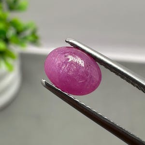 May include: A vibrant, oval-shaped, translucent pink gemstone held by a pair of silver-toned metal tweezers. The stone features a smooth, polished cabochon surface with natural internal inclusions visible under the light, showcasing a deep magenta hue.