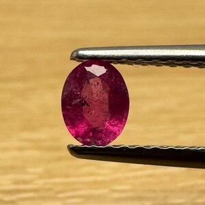 Op de afbeelding: Een gefacetteerde, ovaalvormige robijn edelsteen, vastgehouden door een pincet. De robijn heeft een diepe, levendige rode kleur en de facetten vangen het licht op, waardoor een sprankelend effect ontstaat. De achtergrond is een lichtgekleurd houten oppervlak.