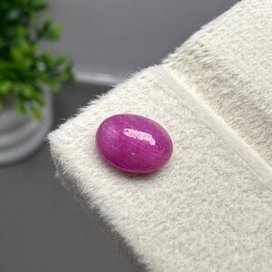 May include: A vibrant, oval-shaped magenta gemstone with a smooth, polished cabochon surface. The stone displays a deep, saturated pinkish-red hue with subtle internal light reflections, resting on a textured, off-white display surface.