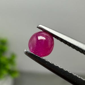 May include: A small, round, vibrant magenta-pink gemstone held by a pair of silver-toned metal tweezers. The stone has a smooth, polished surface with a glossy finish that reflects light, showcasing its deep, saturated color and circular shape.