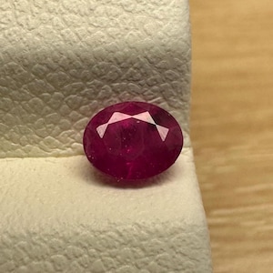 Puede incluir: Gema de rubí ovalada y facetada. El rubí es de color rojo intenso y vibrante y se muestra sobre una superficie texturizada de color blanco roto. La gema se encuentra sobre un fondo de madera.