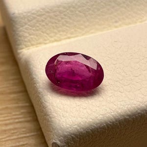 May include: A vibrant, oval-cut ruby gemstone. The ruby, with its deep red hue, is displayed on a white surface. The gemstone's facets catch the light, showcasing its clarity and brilliance. This precious stone is a beautiful example of natural jewelry.