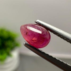 Könnte beinhalten: Ein tropfenförmiger, polierter rosaroter Edelstein, der von einer silberfarbenen Metallpinzette gehalten wird. Der Stein hat eine glatte, abgerundete Oberfläche mit sichtbaren Einschlüssen und einem hellen weißen Lichtreflex auf der Oberseite.