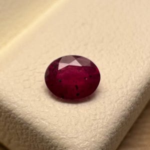 Puede incluir: Un rubí ovalado y facetado, de color rojo intenso, con inclusiones oscuras. La gema se encuentra sobre una superficie texturizada de color crema. El rubí es el punto focal, mostrando su belleza natural y potencial para joyería.