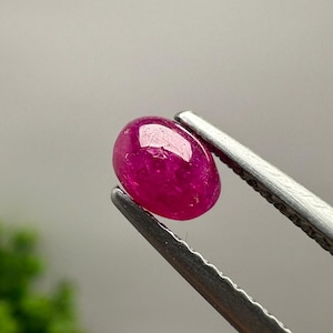 May include: A vibrant, oval-shaped magenta gemstone with a smooth, polished cabochon surface. The stone exhibits a deep, saturated pinkish-red hue with subtle internal inclusions visible under light, held securely by silver-toned metal tweezers.