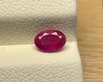 Könnte beinhalten: Ein oval geschliffener Rubin-Edelstein in tiefroter Farbe. Der Rubin liegt auf einer strukturierten, cremefarbenen Oberfläche. Die Facetten des Edelsteins fangen das Licht ein und zeigen seinen Glanz. Im Hintergrund befinden sich eine Holzoberfläche und ein grauer Stoff.
