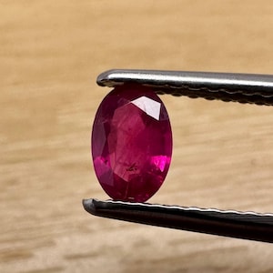 May include: A vibrant, oval-cut ruby gemstone held by tweezers. The ruby displays a deep red hue with facets that catch the light. The tweezers are silver-colored, and the background is a light brown wooden surface.