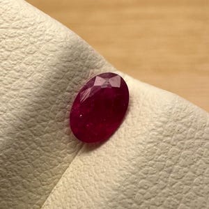 May include: A faceted, oval-shaped ruby gemstone. The ruby is a deep, rich red color and is set against a textured, off-white surface. The gemstone is displayed on a light brown wooden surface.