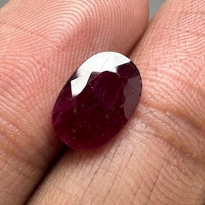 Puede incluir: Una gema de rubí facetada de forma ovalada. El rubí es de un color rojo intenso y rico con una superficie brillante y reflectante. La gema se sostiene en una mano, mostrando su tamaño y forma.