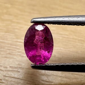 Può includere: Una gemma ovale rosa-rossa vibrante è tenuta da una pinzetta. La gemma ha una superficie sfaccettata, che riflette la luce. La gemma è di un colore intenso e saturo, con alcune inclusioni interne visibili.