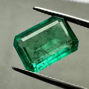 May include: A vivid green emerald gemstone, held by tweezers. The rectangular-cut gem displays internal inclusions and facets. The emerald's shape is rectangular, highlighting its clarity and brilliance. The background is a soft, neutral tone.