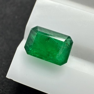 May include: A vivid, emerald-cut emerald gemstone. The gem is a deep, rich green colour and has a rectangular shape with faceted edges. It is displayed on a white surface against a dark background.