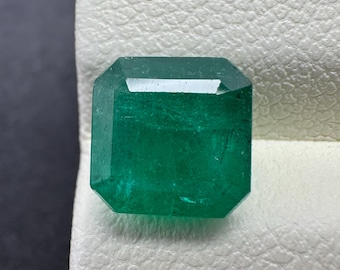 Puede incluir: Una vibrante piedra preciosa verde con talla esmeralda. La gema tiene forma rectangular con bordes biselados, mostrando un color profundo y rico. La piedra está sobre una superficie texturizada blanquecina.
