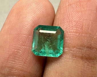 Puede incluir: Una esmeralda rectangular, tallada en esmeralda. La gema es de un color verde intenso y rico, con bordes facetados que reflejan la luz. La esmeralda se muestra entre dos dedos, resaltando su claridad y color.
